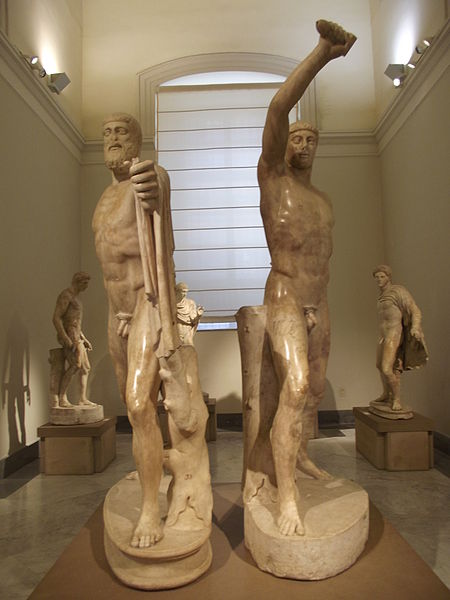 A statue of Harmodius and Aristogeiton in the National Archaeological Museum of Naples. Harmodius has his right arm raised, thrusting his sword forward. Aristogeiton has a cape draped over his left shoulder and is holding a sword in his left hand.